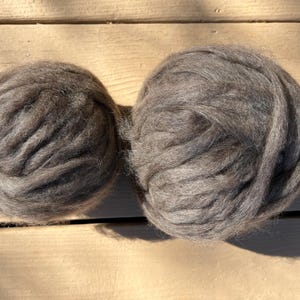 May include: Two gray wool roving balls, ideal for fiber art projects, sit on a wooden surface. The wool has a soft, textured appearance, perfect for needle felting, wet felting, or arm knitting. The natural fibers are a neutral color.