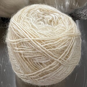 May include: A large ball of off-white yarn, tightly wound. The yarn appears soft and fluffy, with a slightly textured surface. The ball is centered in the frame, with a neutral background.