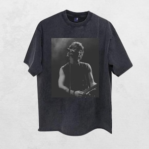May include: Dark grey t-shirt with a black and white image of a musician performing on stage. The image shows the musician playing a guitar and singing. The shirt has a crew neck and short sleeves. The image is centered.
