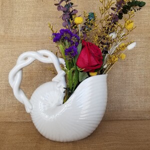 May include: A white ceramic seashell-shaped vase with a twisted handle, filled with a vibrant arrangement of dried flowers. The arrangement includes a red rose, purple statice, yellow billy buttons, and eucalyptus.