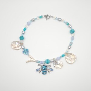 May include: A beaded necklace featuring a silver clasp and a variety of beads in shades of blue, white, and gray. The necklace includes a bee pendant, flower, and shell-shaped charms. The necklace is approximately 18 inches in length.