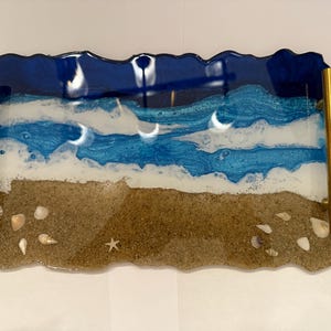 May include: A rectangular resin tray with a beach scene. The tray features a dark blue ocean, white foamy waves, and a sandy beach with seashells and a starfish. Gold handles are attached to the sides.