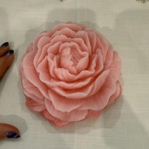 Könnte beinhalten: Eine große, hellrosa, blütenförmige Seife mit detaillierten Blütenblättern. Die Seife befindet sich auf einem weißen Tuch mit Schneeflockenstickerei. Die Farbe der Seife ist ein weiches, pastellfarbenes Rosa.
