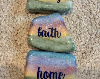 May include: Three smooth, polished stones painted with a gradient of blue, pink, yellow, and green colors. Each stone features dark blue cursive lettering. The top stone reads family, the middle stone reads faith, and the bottom stone reads home.