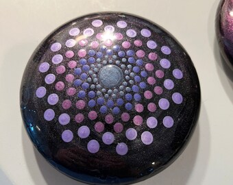 Pode incluir: Um coaster redondo preto com um design de círculo concêntrico em tons de roxo, azul e branco.