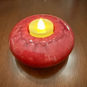 May include: A red, round candle holder with a built-in, battery-operated, yellow LED candle. The candle holder has a glossy finish and a circular indentation to hold the candle. The candle's flame is shaped like a teardrop.