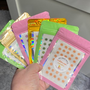 Pode incluir: Quatro sacos lacráveis com fundos de cores diferentes, cada um contendo pequenas aspersões redondas. O saco rosa tem um adesivo que diz "OH SNAP DOTZ" e "maliby rum cupcakes".