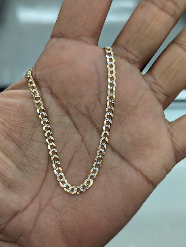 May include: A gold-colored curb chain necklace with alternating gold and silver links. The chain is held in a hand, showcasing its design and texture. The necklace is a jewelry item.