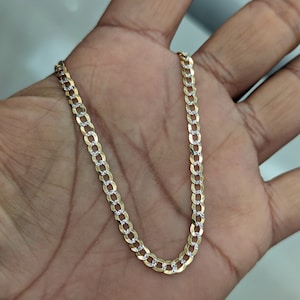 May include: A gold-colored chain necklace with a curb link design. The chain has alternating gold and silver-colored links, creating a two-tone effect. The necklace is displayed on a person's open palm.