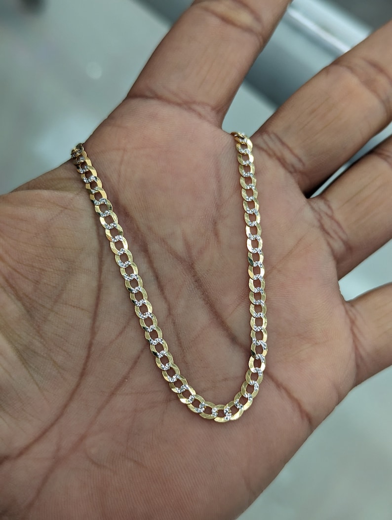May include: A gold-colored chain necklace with a curb link design. The chain has alternating gold and silver-colored links, creating a two-tone effect. The necklace is displayed on a person's open palm.