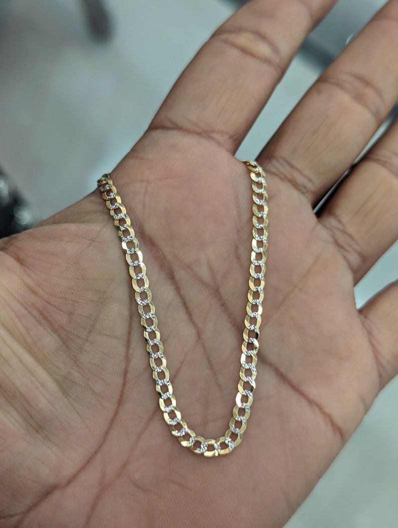 May include: A gold-colored curb chain necklace with alternating textured and smooth links. The chain is displayed on a hand, showcasing its design and texture. The necklace is a jewelry item.
