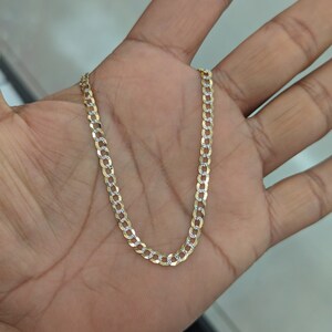 May include: A gold-colored curb chain necklace with alternating gold and silver-colored links. The necklace is displayed on a person's open palm, showcasing its design and detail. The chain is a classic jewelry piece.