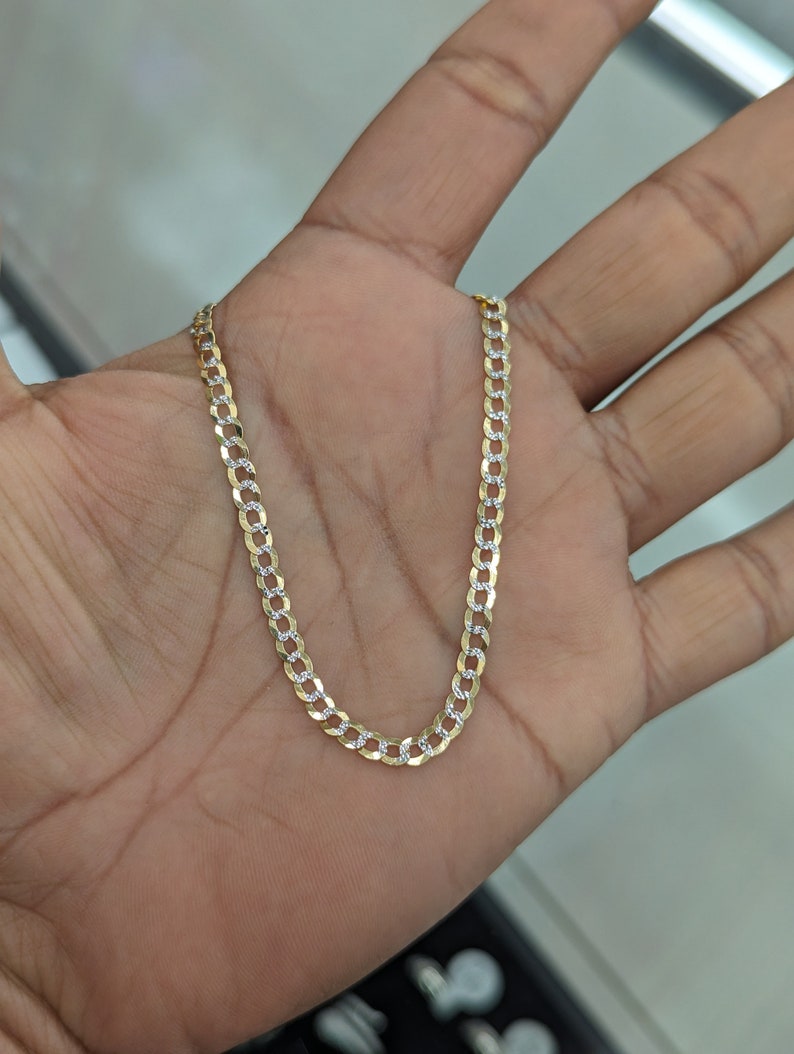 May include: A gold-colored curb chain necklace with alternating gold and silver-colored links. The necklace is displayed on a person's open palm, showcasing its design and detail. The chain is a classic jewelry piece.