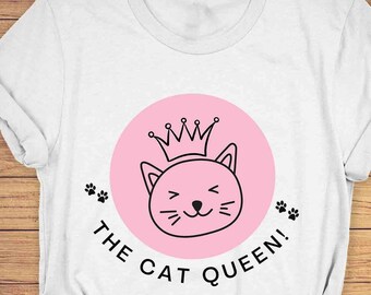 Peut inclure: T-shirt unisexe blanc avec un cercle rose et un dessin au trait noir d'un chat portant une couronne. Le texte "THE CAT QUEEN!" est imprimé sous le cercle.