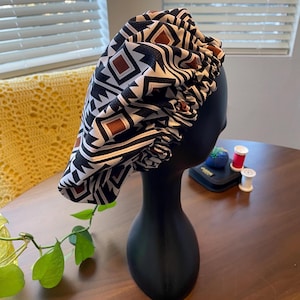 May include: A black satin bonnet with a geometric pattern in white and brown is displayed on a black mannequin head. The bonnet has an elastic band and is designed to protect hair. The background includes a wooden table and a yellow crocheted blanket.