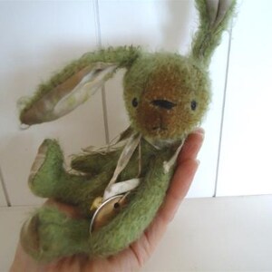 May include: A green and brown plush bunny rabbit toy with a white ribbon around its neck. The bunny has a long, floppy ear and a sweet expression.