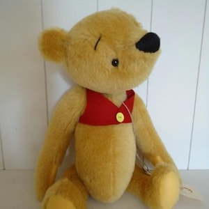 May include: A plush teddy bear with brown fur, a red vest with a yellow button, and a black nose. The bear is sitting on a white surface.
