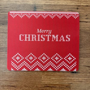 May include: A red Christmas card with a knitted pattern design. The card features the words "Merry CHRISTMAS" in white, with white decorative borders at the top and bottom. The card is rectangular and lies on a wooden surface.