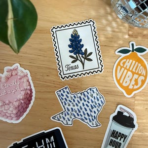 Pu&ograve; includere: Una collezione di adesivi su una superficie di legno. Gli adesivi includono un francobollo Texas bluebonnet, un adesivo a forma di Texas con bluebonnets, un adesivo rosa a forma di cuore, un adesivo con uno shaker per cocktail che dice "Happy Hour", un adesivo con un limone che dice "Chilton Vibes" e un orologio digitale che dice "6:44 AM TIME".