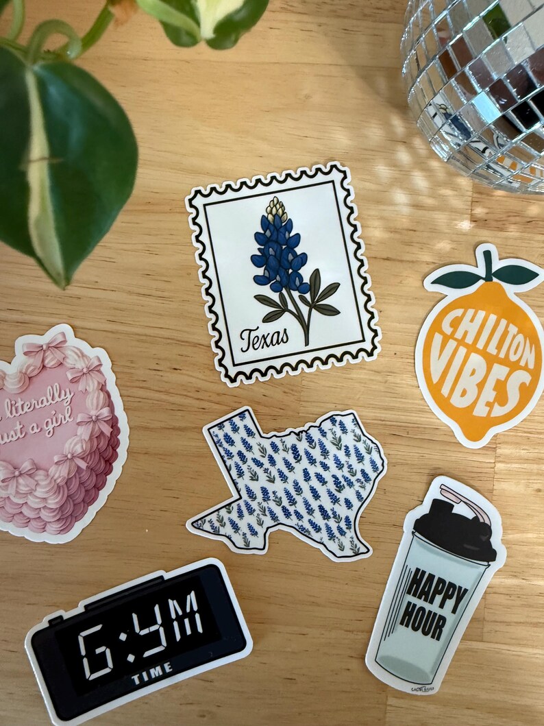 Pu&ograve; includere: Una collezione di adesivi su una superficie di legno. Gli adesivi includono un francobollo Texas bluebonnet, un adesivo a forma di Texas con bluebonnets, un adesivo rosa a forma di cuore, un adesivo con uno shaker per cocktail che dice "Happy Hour", un adesivo con un limone che dice "Chilton Vibes" e un orologio digitale che dice "6:44 AM TIME".