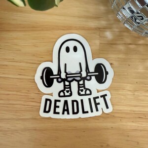 Pu&ograve; includere: Un adesivo bianco con un fantasma dei cartoni animati che solleva un bilanciere. Il fantasma ha due occhi neri e indossa calzini e scarpe a forma di cuore. La parola "DEADLIFT" &egrave; stampata in grassetto, in lettere nere sotto.