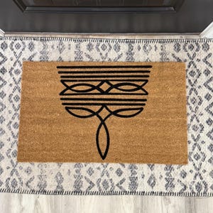 May include: A rectangular coir doormat with a black geometric design. The mat is a natural brown color and features a symmetrical pattern of lines and shapes. The doormat is placed on a patterned rug with a white and gray design.