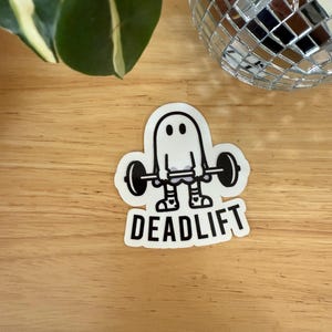 Pu&ograve; includere: Un adesivo bianco con un fantasma dei cartoni animati che solleva un bilanciere. Il fantasma ha due occhi neri e indossa scarpe da ginnastica. La parola "DEADLIFT" &egrave; stampata in grassetto, in nero, sotto.