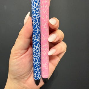 May include: Two ballpoint pens, one blue with white floral pattern, the other pink with a similar design. Both pens have silver-colored tips and black ink tips. The pens are held against a dark gray background.