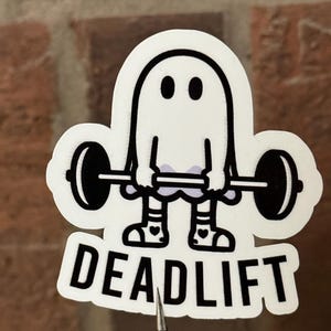 Pu&ograve; includere: Un adesivo bianco con un fantasma dei cartoni animati che solleva un bilanciere. Il fantasma ha occhi neri e indossa scarpe con disegni a cuore. La parola "DEADLIFT" &egrave; stampata in grassetto nero sotto.