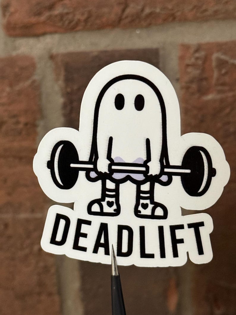Pu&ograve; includere: Un adesivo bianco con un fantasma dei cartoni animati che solleva un bilanciere. Il fantasma ha occhi neri e indossa scarpe con disegni a cuore. La parola "DEADLIFT" &egrave; stampata in grassetto nero sotto.
