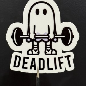 Pu&ograve; includere: Un adesivo bianco con un fantasma dei cartoni animati che solleva un bilanciere. Il fantasma ha due occhi neri e indossa calzini a righe e scarpe con cuori. La parola "DEADLIFT" &egrave; stampata in grassetto, in lettere nere.