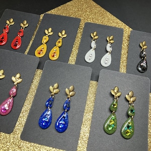 Peut inclure: Assortiment de boucles d'oreilles pendantes avec des attaches dorées et des gouttes de résine en forme de larme. Les boucles d'oreilles sont décorées de paillettes et de strass de différentes couleurs, notamment rouge, jaune, rose, bleu, blanc, noir et vert.