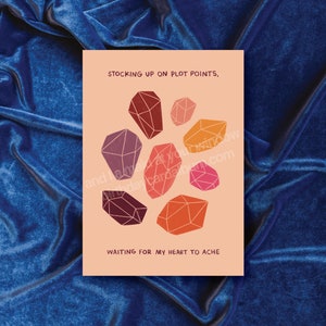 May include: A peach-coloured card with the text "STOCKING UP ON PLOT POINTS, WAITING FOR MY HEART TO ACHE" and a design of seven colourful geometric shapes resembling gemstones.