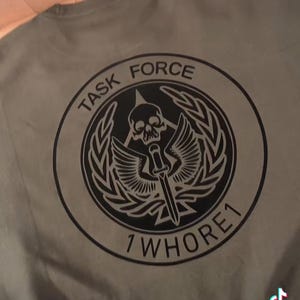 May include: A green t-shirt with a black circular graphic. The graphic features a skull with wings, a dagger, and a laurel wreath. The text "TASK FORCE 1 WHORE 1" is printed around the graphic.