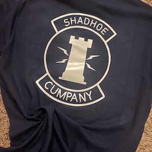 Puede incluir: Una camiseta azul marino con un gráfico blanco de un castillo y el texto "SHADHOE COMPANY" en la espalda.