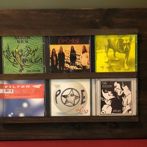 May include: A wooden CD rack with six slots, displaying five CDs. The CDs feature various artists and album titles, including "Saigon Kick", "Candlebox", "Filter", "Poe", and "Mad Season".