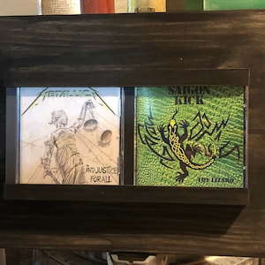 Può includere: Un ripiano in legno scuro per CD con due custodie per CD. La custodia a sinistra presenta un'immagine in bianco e nero di una donna che tiene una bilancia con il testo "And Justice For All". La custodia a destra ha uno sfondo verde con una lucertola nera e il testo "Saigon Kick The Lizard".