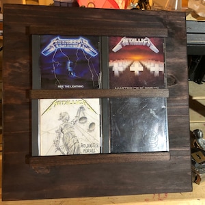 May include: A dark stained wooden CD display case holding four Metallica albums. The albums are "Ride the Lightning", "Master of Puppets", "...And Justice for All", and one other. The case has a square frame with horizontal supports.