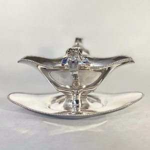 Könnte beinhalten: Eine silberne Sauciere mit passender Untertasse. Das Boot hat einen dekorativen Griff und einen gewellten Rand. Das Set besteht aus glänzendem, reflektierendem Metall, wahrscheinlich Silber oder versilbertem Material. Das Design ist aufwendig und elegant.