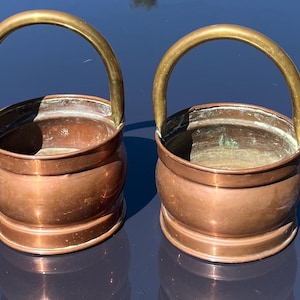 May include: Two copper buckets with brass handles. The buckets have a rounded body and a wide rim. The handles are arched and attached to the sides. The copper has a warm, reddish-brown colour.