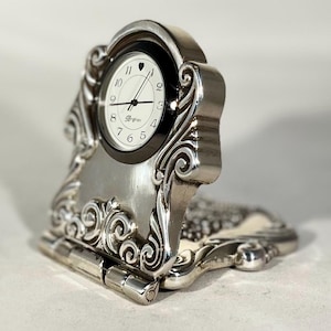 May include: Silver-coloured, hinged desk clock with an ornate, scrolled design. The clock face is white with black numerals and hands, and the brand name "Brighton" is visible. The clock is set against a neutral background.