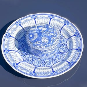 Puede incluir: Un plato y un recipiente con tapa de cerámica azul y blanca con un diseño de calendario, meses, días e ilustraciones celestiales que incluyen sol, luna y planetas. El texto "CELEBRATE THE MILLENNIUM" aparece alrededor del recipiente.