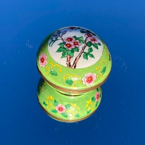 Puede incluir: Una pequeña caja redonda de color verde con un diseño floral. La tapa tiene un fondo blanco con flores rosas y hojas verdes, mientras que la base es verde con detalles amarillos.