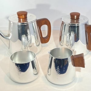 Può includere: Un set da tè e caffè di quattro pezzi con corpi in metallo color argento lucido e manici e pomelli del coperchio in legno marrone scuro. Il set include una teiera grande, una caffettiera più piccola, una lattiera e una zuccheriera, ciascuna con un motivo a foglia.