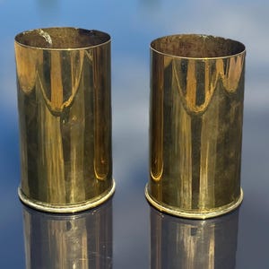 May include: Two cylindrical brass vessels with a polished, reflective gold-toned finish. Each container features a smooth, straight-walled body and a slightly flared base rim, presenting a uniform metallic appearance with visible surface reflections.