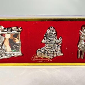 Può includere: Un set di ornamenti natalizi argentati in una scatola di velluto rosso con finiture dorate. Il set include una cornice per foto con un cartello del Polo Nord, una figura di Babbo Natale e una renna. La scatola è rettangolare.