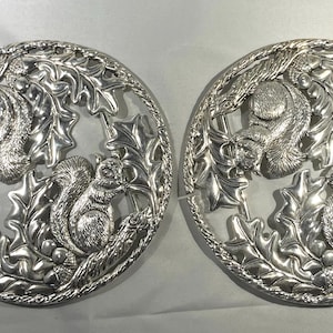 Könnte beinhalten: Zwei silberfarbene, kreisförmige Dekorationsstücke. Jedes zeigt ein detailliertes Relief von Eichhörnchen inmitten von Eichenblättern und Eicheln. Das Design wird von einem geflochtenen Rand umrahmt, wodurch eine von der Natur inspirierte Ästhetik entsteht.