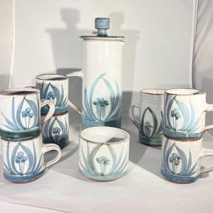 May include: A ceramic coffee press and matching mugs. The set features a white background with blue floral designs. The mugs have handles and are stacked. The coffee press has a lid and a handle. The set is handmade and has a rustic aesthetic.