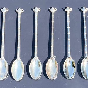 Könnte beinhalten: Ein Set aus sechs silberfarbenen Metalllöffeln, die in einer Reihe angeordnet sind. Jeder Löffel hat einen langen, schlanken Stiel mit segmentiertem Design und einen kleinen, dekorativen Aufsatz in Teekannenform. Die Löffel haben ovale Laffen.