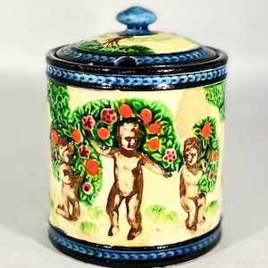 May include: A ceramic jar with a lid, depicting cherubic figures in a garden with fruit trees. The jar has a blue rim and a blue knob on the lid. The background is a light yellow colour. The jar is approximately 10 cm tall.
